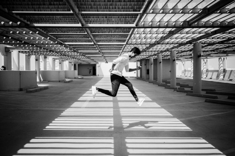 Person jumping mid-air in a sunlit parking garage, shadows forming lines on the ground.