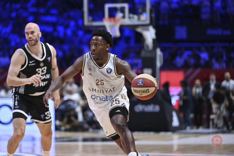 A basketball player in a white jersey dribbles past a defender in a black jersey during a game.