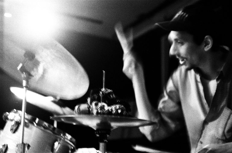 A drummer plays passionately on a drum set under bright lighting in a black and white photo.