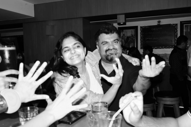 Two people smiling and posing with outstretched hands at a busy bar, surrounded by friends and drinks.