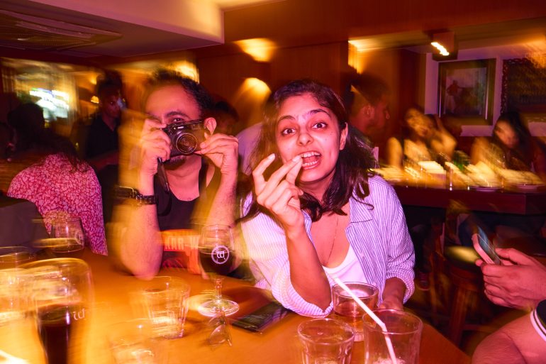 Woman gesturing at camera and man taking a photo in a lively, colorful, and blurry restaurant scene.