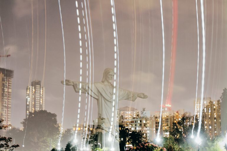 Long-exposure photo of a Christ statue with city buildings and streaks of light in the night sky.