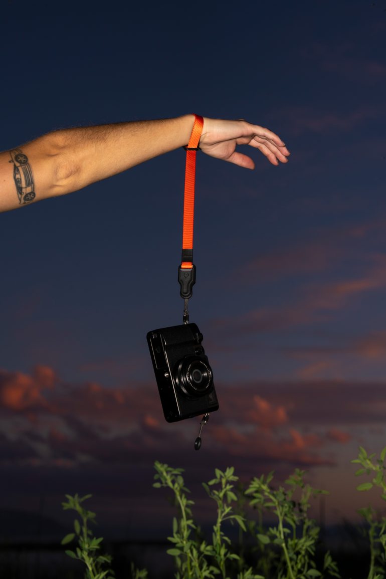 An arm with a tattoo holds a camera by an orange strap against a sunset sky and green plants.