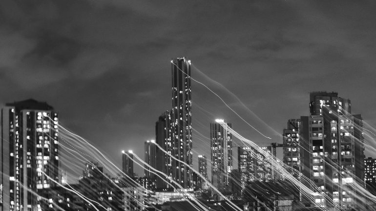 Black and white city skyline at night with light trails creating streaks across the tall buildings.