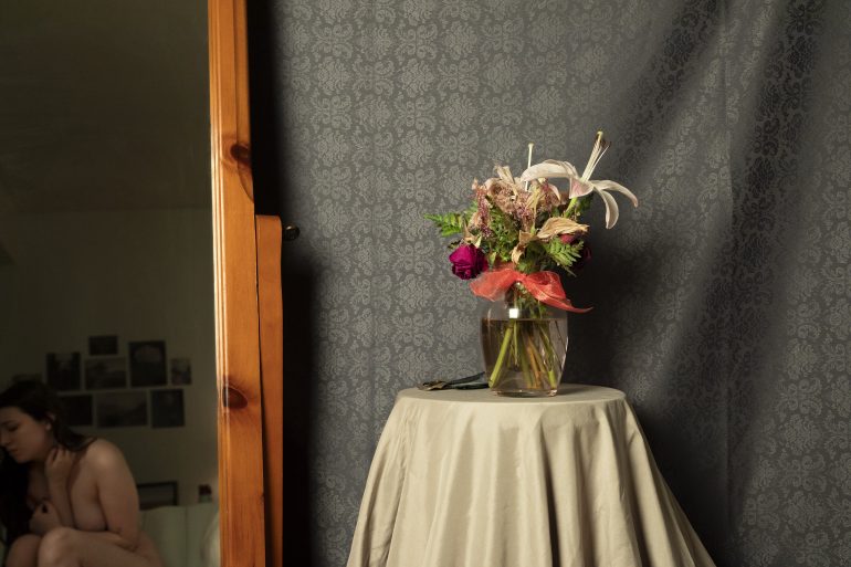 A vase of flowers on a draped table, with a woman sitting in the background near a mirror.