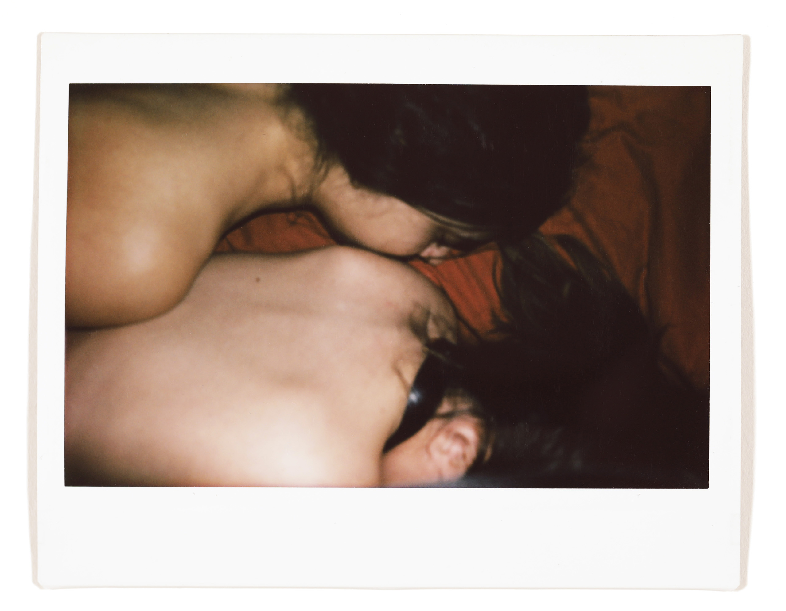 Two people lying close together on a bed, both are topless and facing away from the camera.