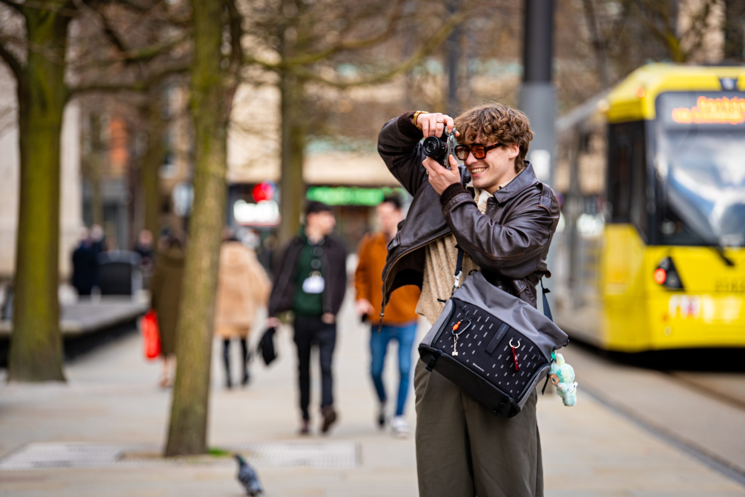 Manfrotto Just Redesigned the Camera Bag for the Modern Photographers A person wearing sunglasses takes a photo on a city street with a yellow tram and pedestrians in the background.