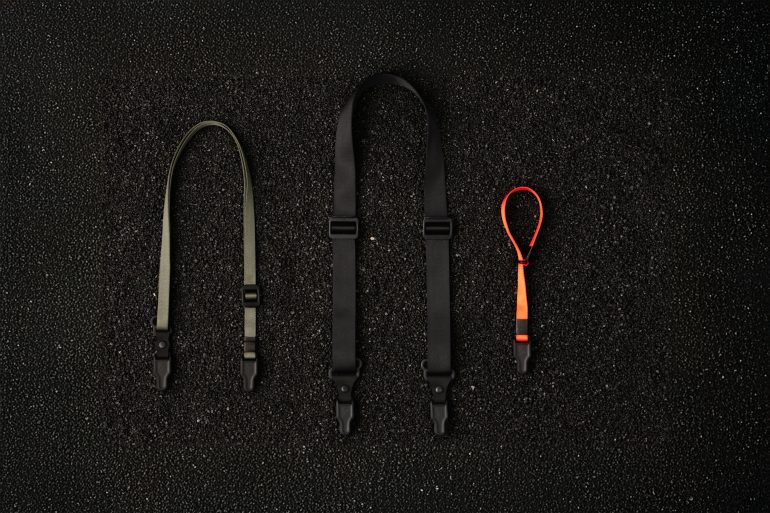 Three different camera straps—short black, long black, and small red—are arranged on a textured black surface.