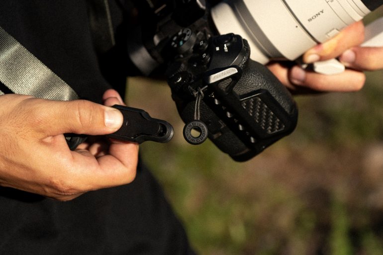 Person attaching a quick-release clip to a black camera with a white lens outdoors in sunlight.
