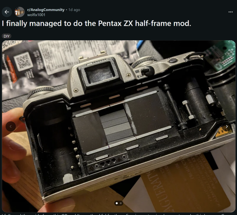 Open Pentax ZX film camera showing an exposed shutter curtain, being held for modification on a cluttered desk.