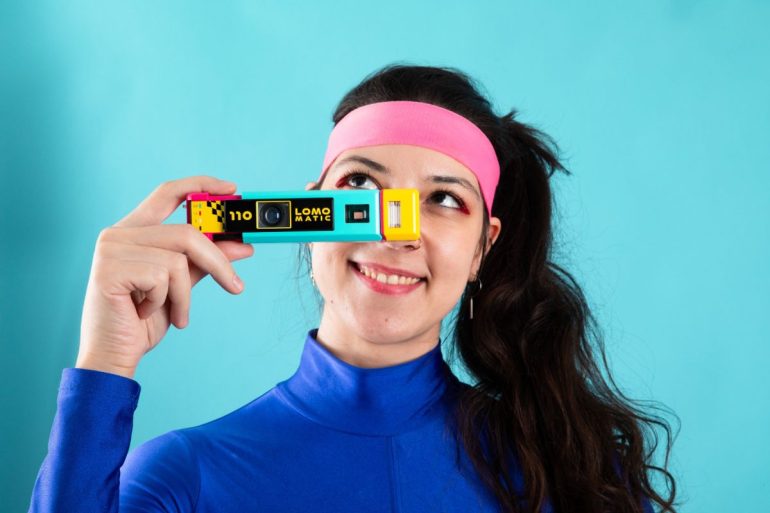 Woman in a blue top and pink headband holding a colorful camera up to her eye, smiling.