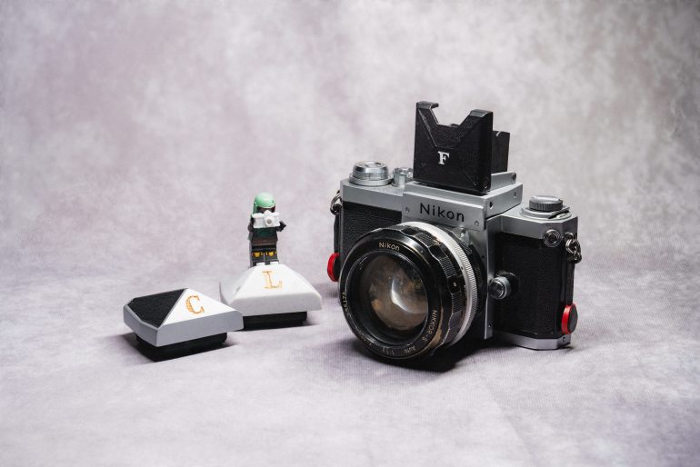 A vintage Nikon camera with lens sits beside two camera parts and a small Lego figure on a gray surface.