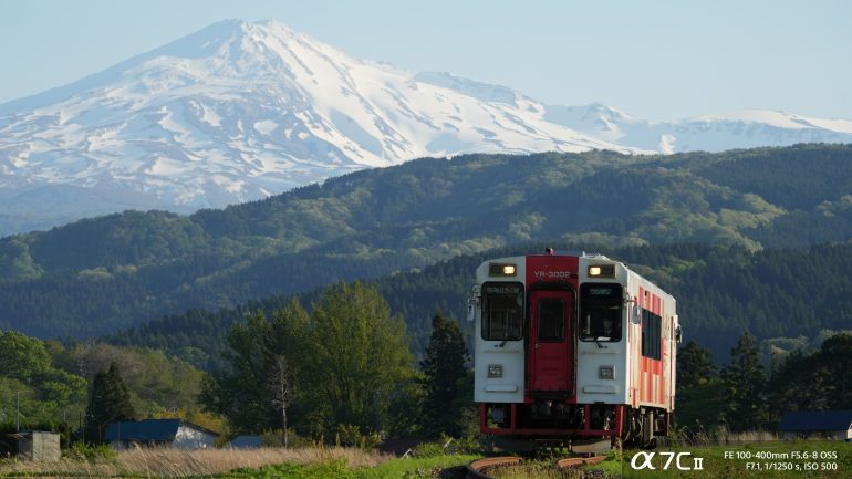 A red and white train travels through greenery with a snow-capped mountain in the background.