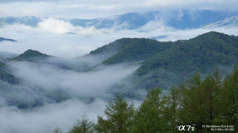 Green mountains covered in mist and clouds, with distant peaks and a partly cloudy sky in the background.