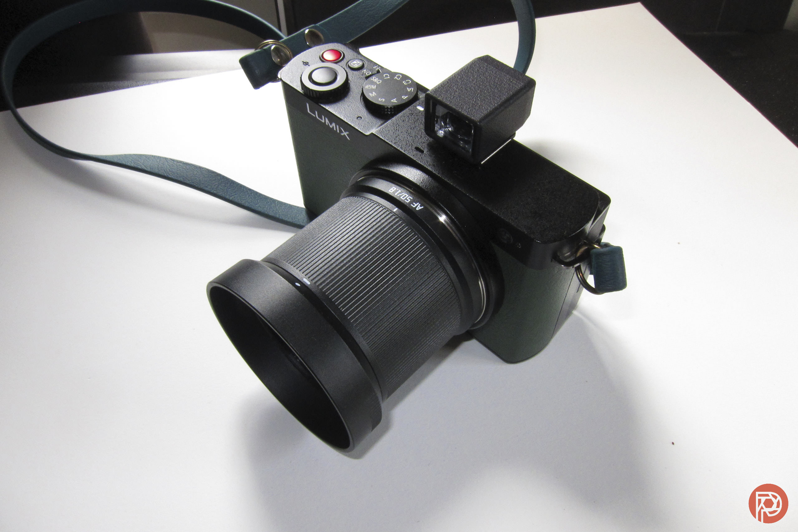 Black Lumix digital camera with large lens and blue strap resting on a white surface.