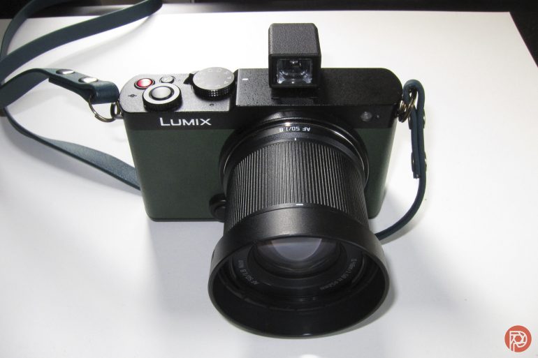 A black Lumix digital camera with a lens and strap, placed on a white surface.