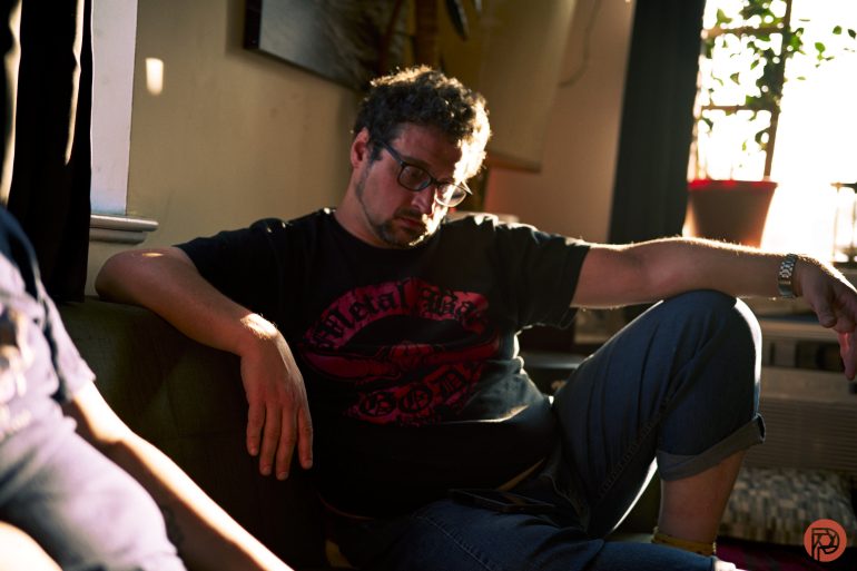 Man with glasses slouches on a couch in sunlight, wearing a black t-shirt and jeans.