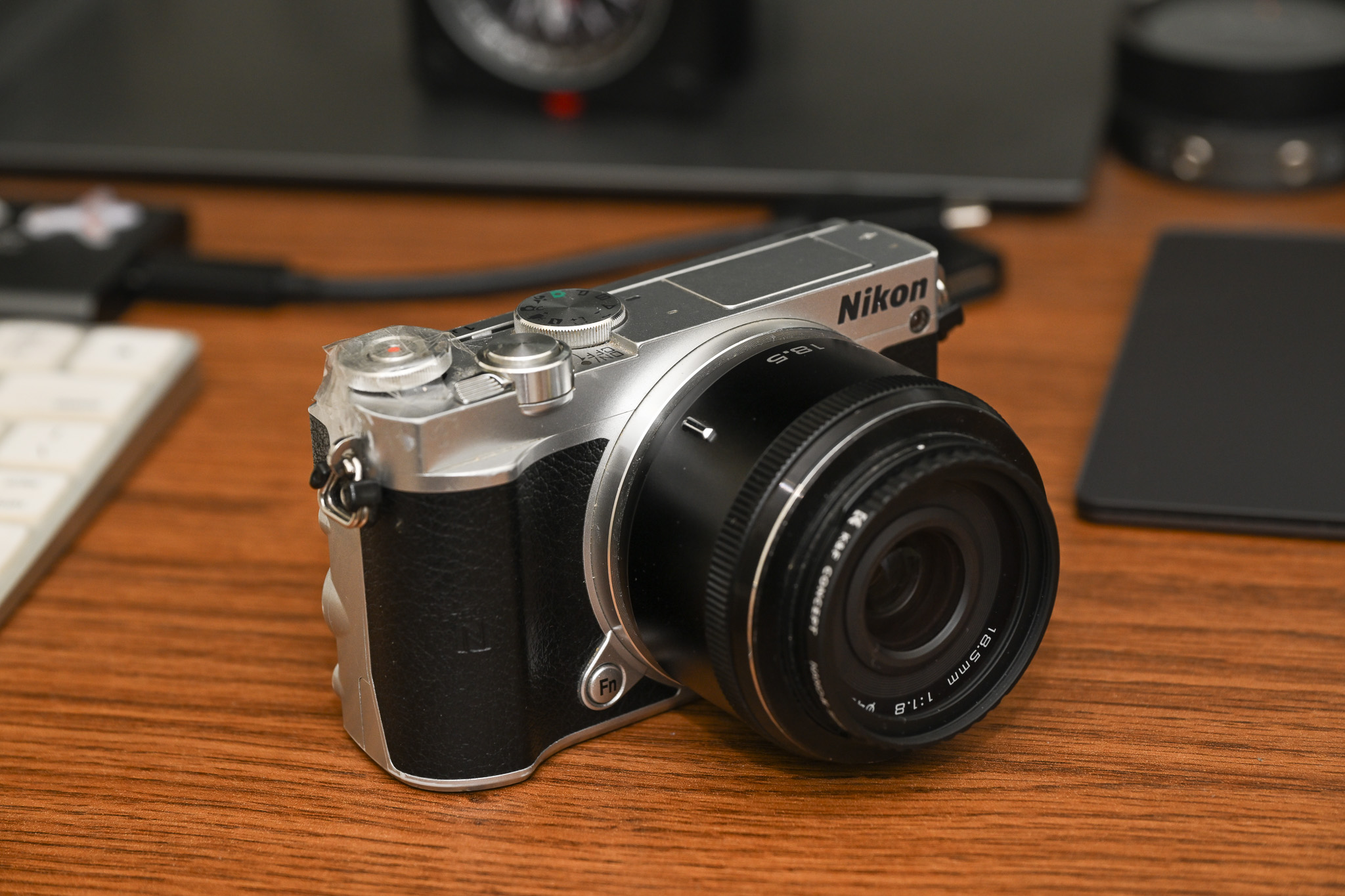 A silver Nikon mirrorless camera with a lens sits on a wooden desk near a keyboard and other items.