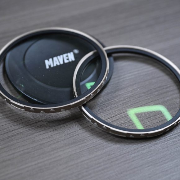 Two camera lens filters and a Maven lens cap on a gray wooden surface, with green reflections on the glass.