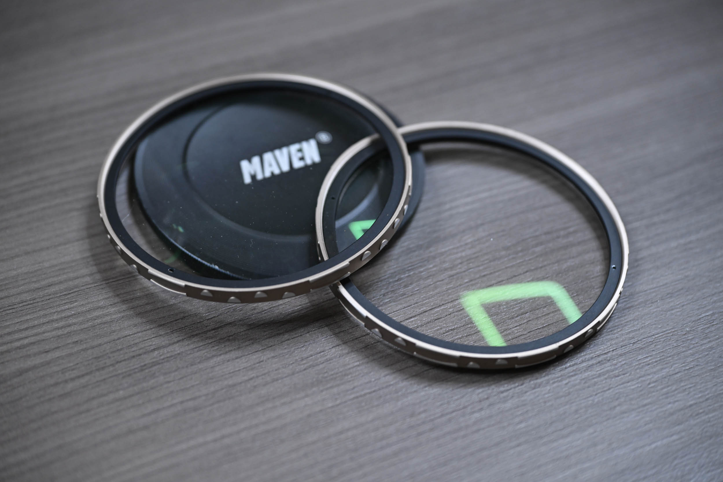 Two camera lens filters and a Maven lens cap on a gray wooden surface, with green reflections on the glass.