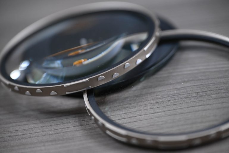 Two metal-rimmed camera lens filters overlap on a wooden surface, one tilted on top of the other.