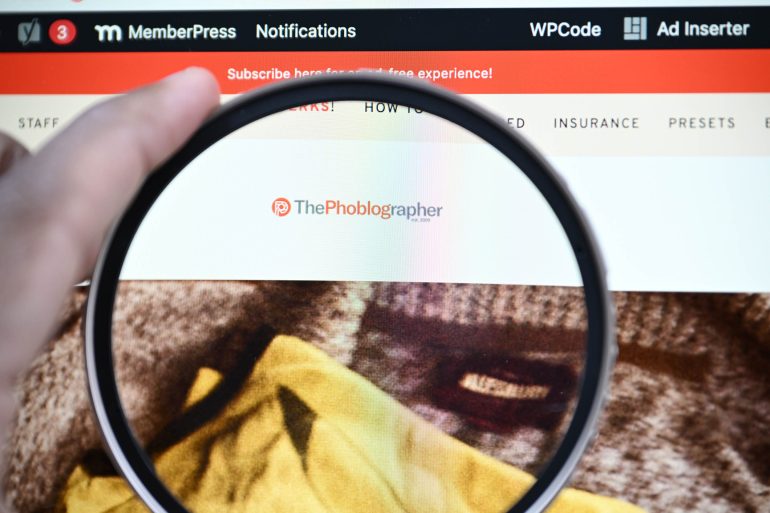 A hand holds a magnifying glass over The Phoblographer website logo on a computer screen.