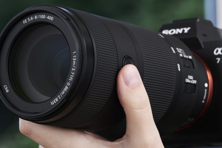 Close-up of a hand holding a Sony Alpha camera with a large 100-400mm zoom lens attached.