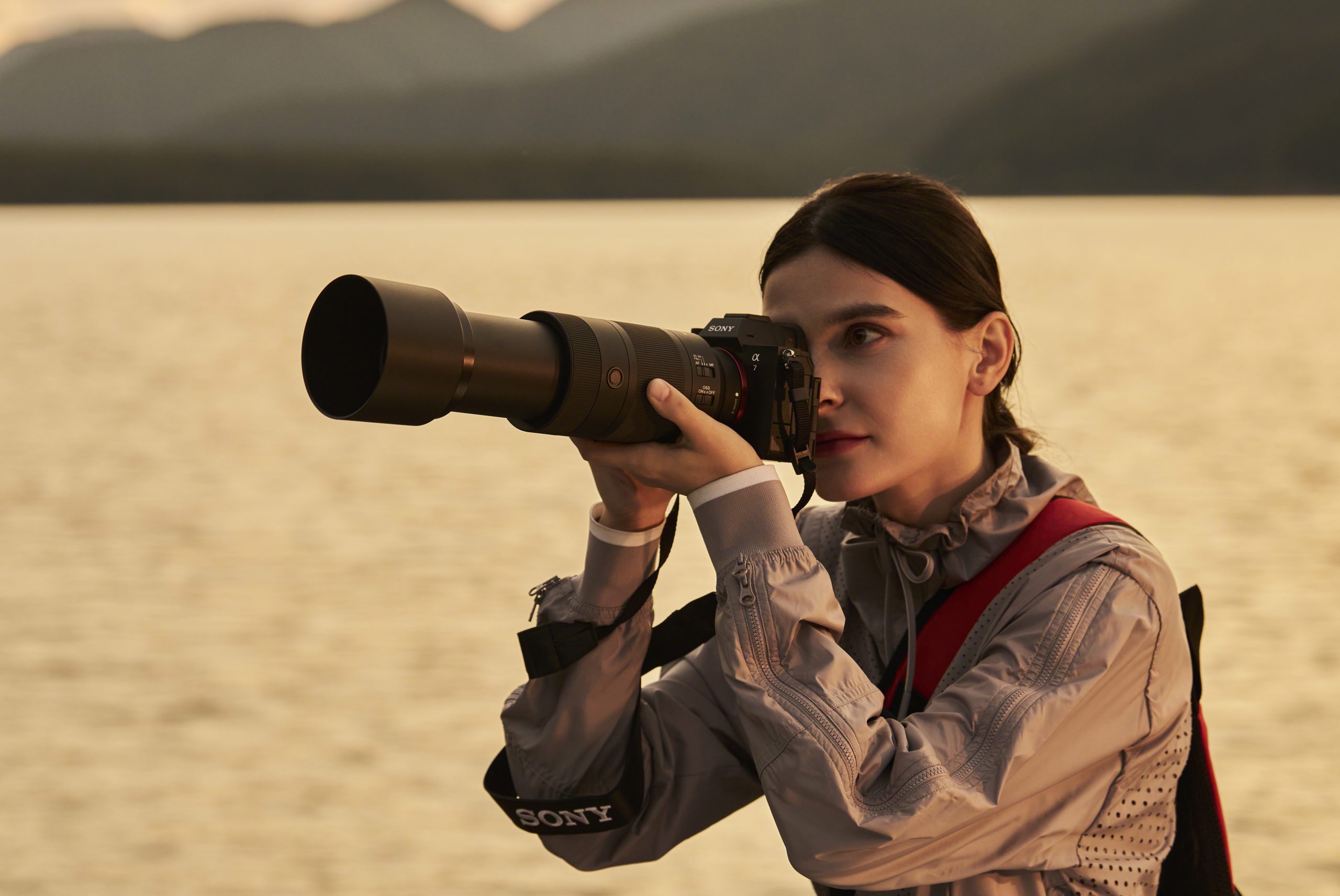 Woman with dark hair using a Sony camera and telephoto lens by a lake at sunset.