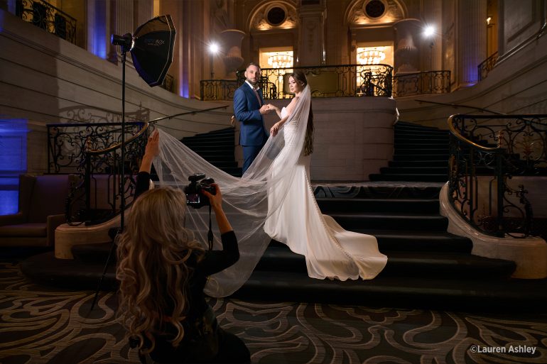 A photographer captures a bride and groom posing on grand stairs in an elegant indoor setting.